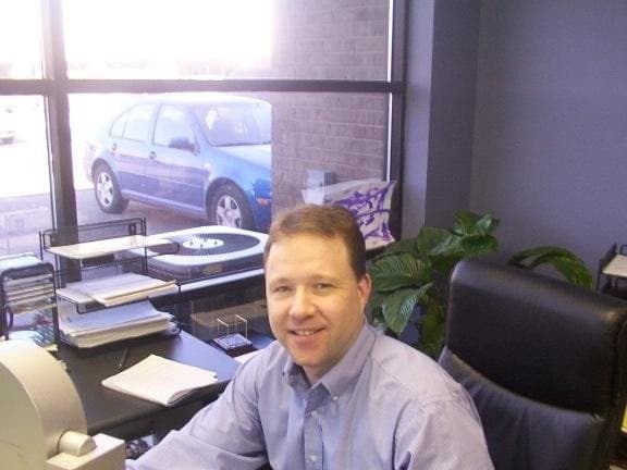 Peter Munro Roberts smiling at his office desk in a blue dress shirt, with paperwork, an office plant, and a car visible through the window behind him.