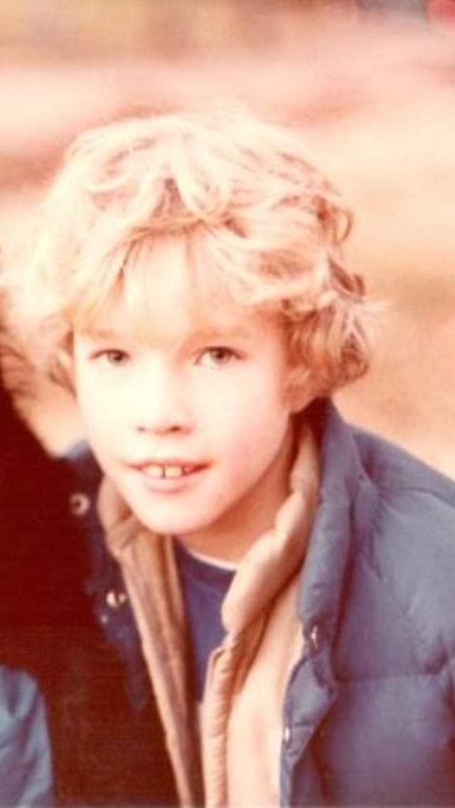 A 1970s-era photograph of a smiling blond boy in a blue puffer jacket outdoors.
