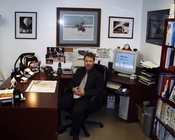 Peter Munro Roberts in a dark suit seated at an L-shaped wooden desk in a compact office, with a CRT computer monitor, desk phone, bookshelf, and framed pictures of a hot-air balloon, a horseback rider, and a locomotive on the walls.