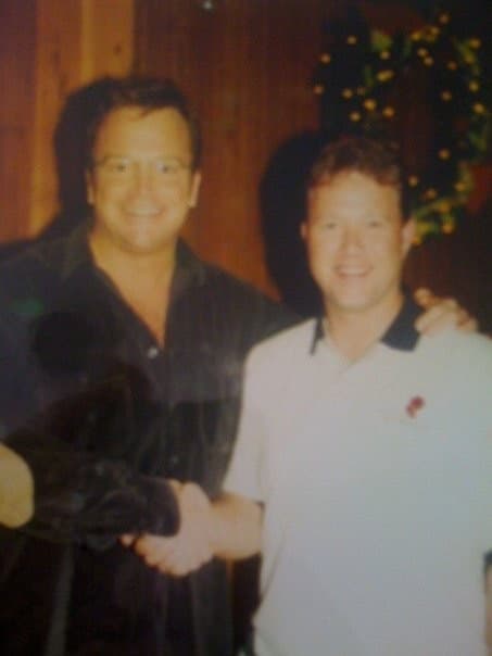 Peter Munro Roberts in a white polo shirt shaking hands with actor Tom Arnold in front of a wood-paneled wall with holiday greenery.